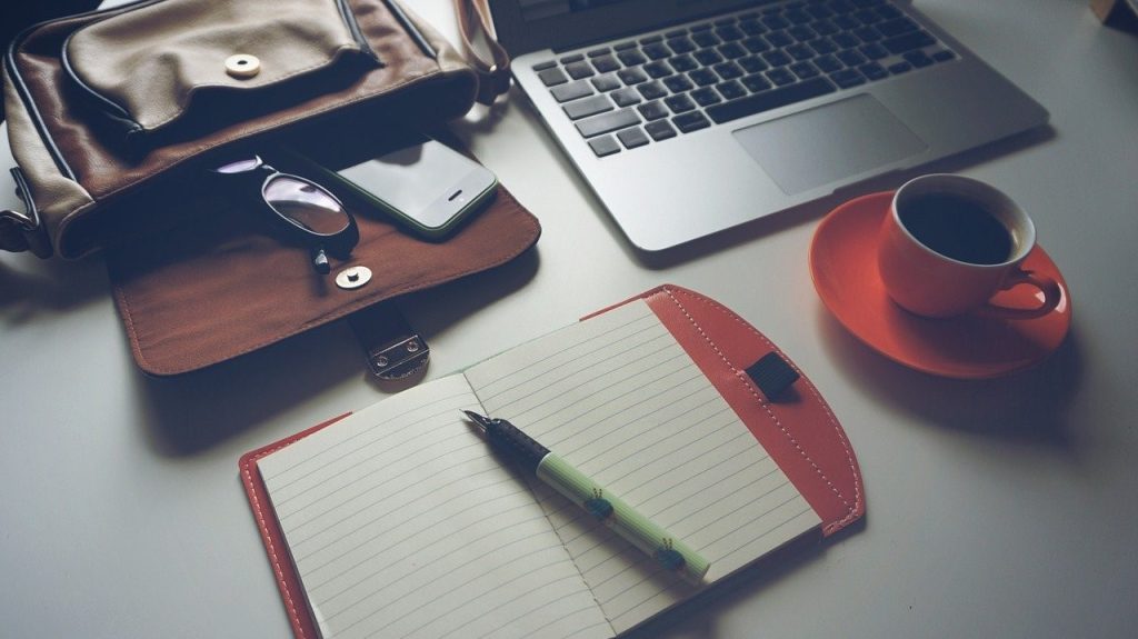 Zaken als een rugzak, laptop, telefoon, bril, notitieboekje met vulpen en een kopje koffie.