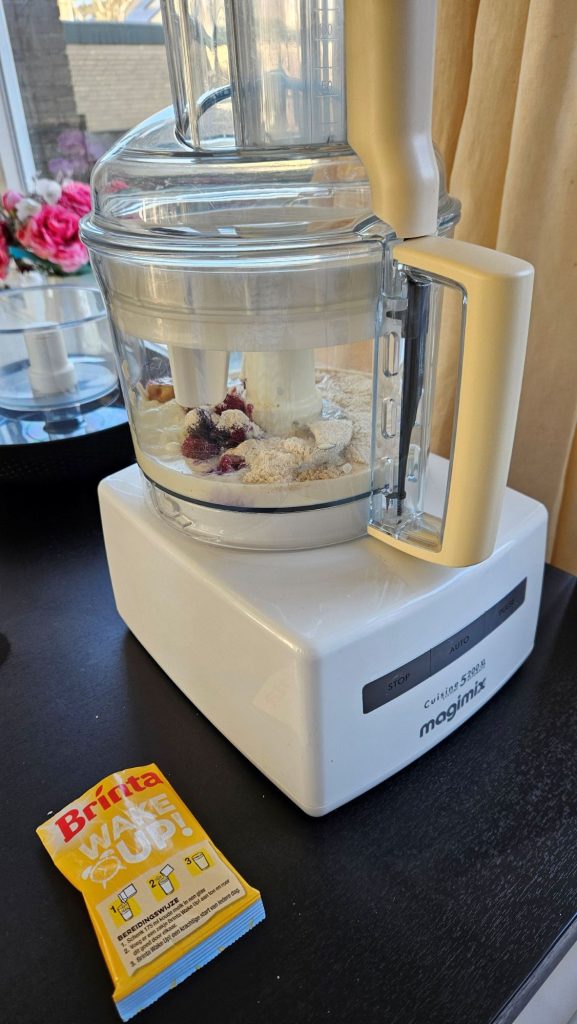 Alle ingrediënten in de foodprocessor met er nat het lege zakje van de Brinta.